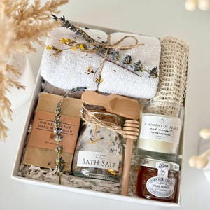 Puede incluir: Una caja de regalo blanca que contiene un set de spa. El set incluye una toalla blanca, jab&oacute;n, sales de ba&ntilde;o, una vela, miel y un cuchar&oacute;n de miel de madera. La caja est&aacute; decorada con flores secas y el texto "Set 1".