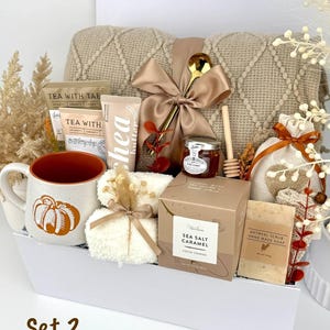 May include: A gift set containing a beige knit blanket with a satin bow, a pumpkin-print mug, tea packets, shea butter, a jar of honey with a wooden dipper, a sea salt caramel cocoa cookie box, oatmeal soap, and a small white towel in a white box.