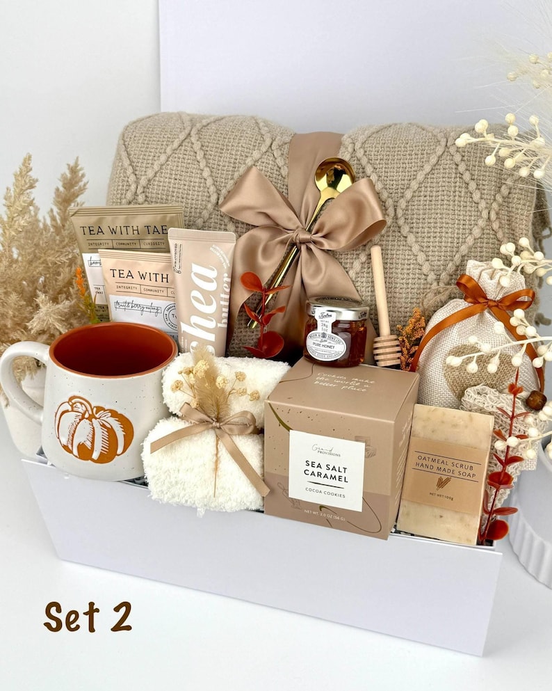 May include: A gift set containing a beige knit blanket with a satin bow, a pumpkin-print mug, tea packets, shea butter, a jar of honey with a wooden dipper, a sea salt caramel cocoa cookie box, oatmeal soap, and a small white towel in a white box.