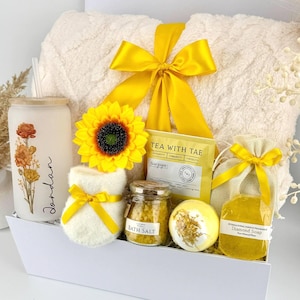 May include: A gift set in a white box containing a cream-colored plush blanket with a yellow ribbon, a frosted glass tumbler with floral art and the name Jordan, a sunflower, a tea packet, bath salts, a bath bomb, and a yellow soap in a pouch.
