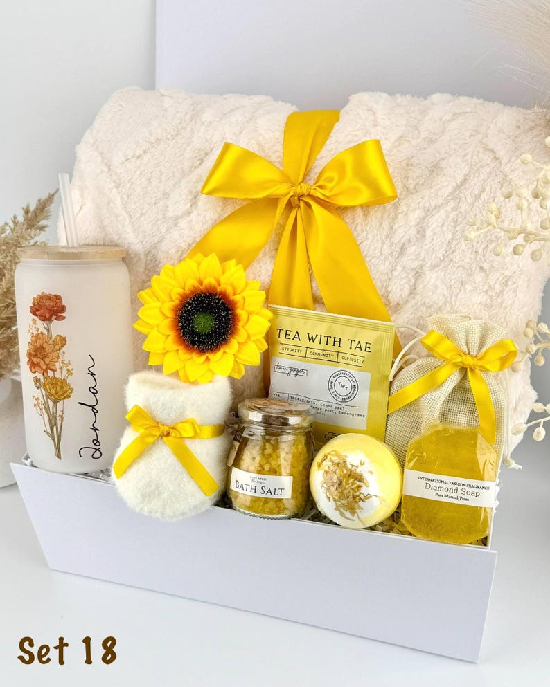 May include: A gift set in a white box containing a cream-colored plush blanket with a yellow ribbon, a frosted glass tumbler with floral art and the name Jordan, a sunflower, a tea packet, bath salts, a bath bomb, and a yellow soap in a pouch.