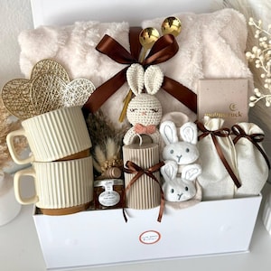 K&ouml;nnte beinhalten: Eine wei&szlig;e Geschenkbox gef&uuml;llt mit einer flauschigen Decke, zwei cremefarbenen Tassen, einem H&auml;schen aus H&auml;kelarbeit, Kerzen und kleinen Geschenkt&uuml;ten. Die Box enth&auml;lt auch Honig, goldene L&ouml;ffel und dekorative Herzen. Der Text "Set 17" ist sichtbar.