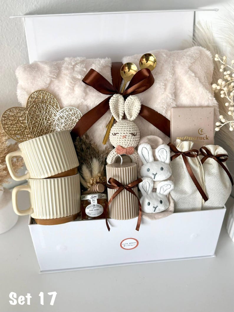 K&ouml;nnte beinhalten: Eine wei&szlig;e Geschenkbox gef&uuml;llt mit einer flauschigen Decke, zwei cremefarbenen Tassen, einem H&auml;schen aus H&auml;kelarbeit, Kerzen und kleinen Geschenkt&uuml;ten. Die Box enth&auml;lt auch Honig, goldene L&ouml;ffel und dekorative Herzen. Der Text "Set 17" ist sichtbar.