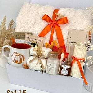 May include: A gift set in a white box containing a cream-coloured plush blanket tied with an orange ribbon, a mug with a pumpkin graphic, two tea packets, a gold spoon, a jar of bath salts, a small jar of honey with a dipper, and a ribbed pillar candle.
