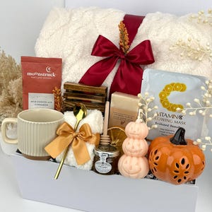 May include: A gift set containing a cream-coloured ribbed mug, a Moonstruck milk chocolate cinnamon bar, a gold spoon, a white fuzzy cloth, a wooden honey dipper, a jar of honey, oatmeal soap, a Vitamin C mask, and a pumpkin-shaped candle.