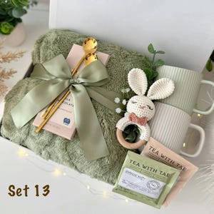 May include: A gift set containing a sage green plush blanket, a milk chocolate bar wrapped in a sage green ribbon with a gold spoon, a cream-colored crocheted bunny rattle, two ribbed mugs in sage and cream, and two Tea With Tae tea packets.