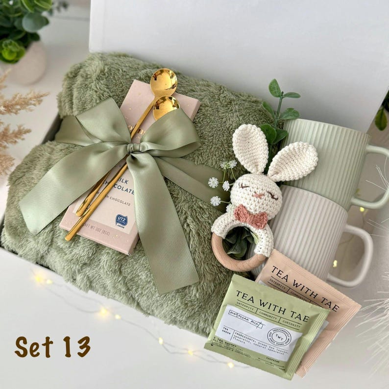 May include: A gift set containing a sage green plush blanket, a milk chocolate bar wrapped in a sage green ribbon with a gold spoon, a cream-colored crocheted bunny rattle, two ribbed mugs in sage and cream, and two Tea With Tae tea packets.