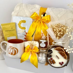 May include: A gift set containing a cream-colored textured blanket with a yellow ribbon, a Moonstruck chocolate bar, a Vitamin C face mask, a pumpkin-patterned mug, a small jar of honey, bath salts, a candle in a coconut shell, and dried flowers.