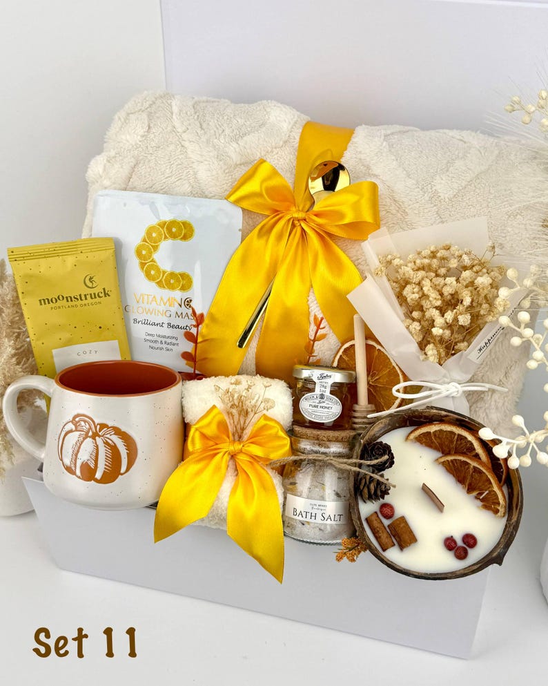 May include: A gift set containing a cream-colored textured blanket with a yellow ribbon, a Moonstruck chocolate bar, a Vitamin C face mask, a pumpkin-patterned mug, a small jar of honey, bath salts, a candle in a coconut shell, and dried flowers.
