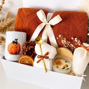 May include: A white gift box containing a folded burnt orange knit blanket tied with a cream ribbon, a white mug with a pumpkin illustration, a gold spoon, a white towel, a candle with a wooden lid, a dried orange slice, and a small pouch.