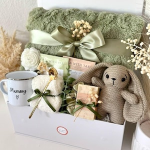 K&ouml;nnte beinhalten: Eine wei&szlig;e Geschenkbox mit einer gr&uuml;nen Decke, Teebeuteln, einer Tasse mit der Aufschrift "Mommy", einem H&auml;schen, Seife und einem goldenen L&ouml;ffel. Die Artikel sind mit gr&uuml;nen B&auml;ndern gebunden. Der Text "Set 14" befindet sich unten.