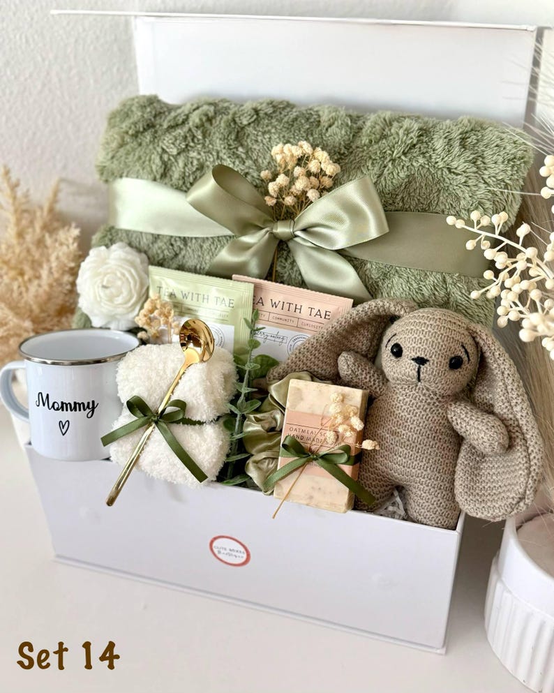 K&ouml;nnte beinhalten: Eine wei&szlig;e Geschenkbox mit einer gr&uuml;nen Decke, Teebeuteln, einer Tasse mit der Aufschrift "Mommy", einem H&auml;schen, Seife und einem goldenen L&ouml;ffel. Die Artikel sind mit gr&uuml;nen B&auml;ndern gebunden. Der Text "Set 14" befindet sich unten.