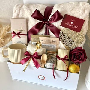 Puede incluir: Una caja de regalo blanca llena de art&iacute;culos de cuidado personal. Incluye una taza color crema, una manta afelpada atada con una cinta burdeos, una barra de chocolate, bolsitas de t&eacute;, sales de ba&ntilde;o, una vela, un coraz&oacute;n decorativo y una rosa roja. La caja est&aacute; etiquetada "Set 16".
