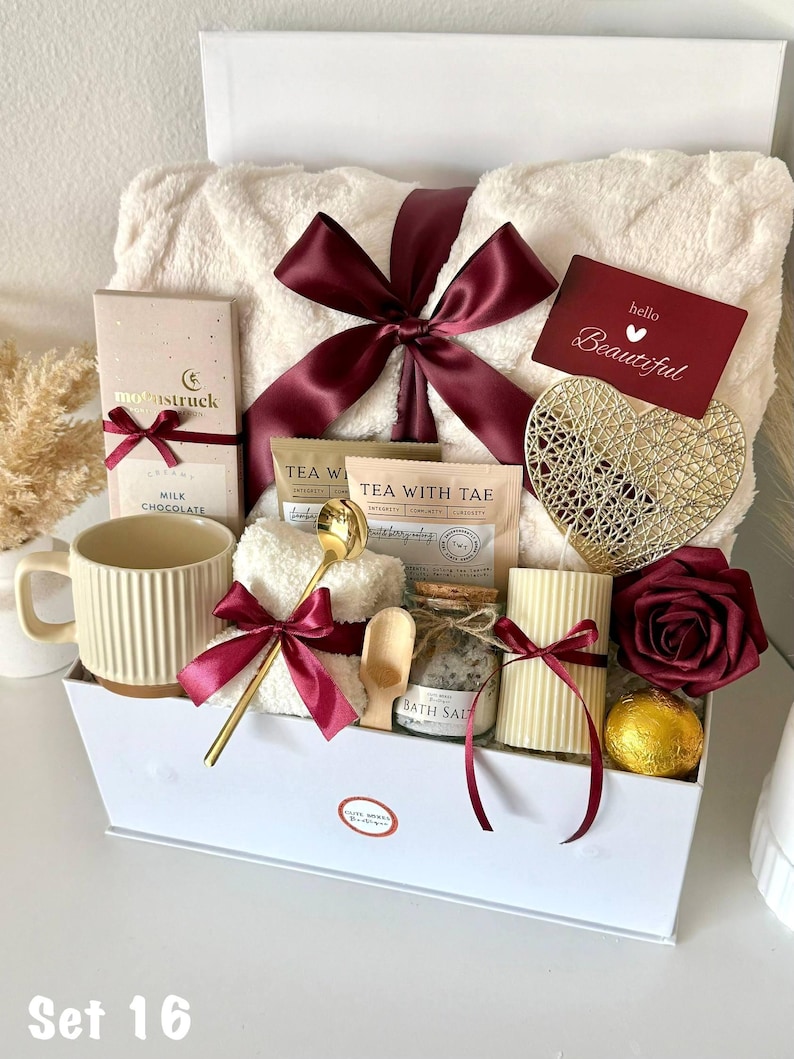 Puede incluir: Una caja de regalo blanca llena de art&iacute;culos de cuidado personal. Incluye una taza color crema, una manta afelpada atada con una cinta burdeos, una barra de chocolate, bolsitas de t&eacute;, sales de ba&ntilde;o, una vela, un coraz&oacute;n decorativo y una rosa roja. La caja est&aacute; etiquetada "Set 16".