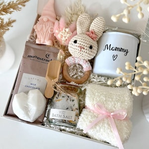 K&ouml;nnte beinhalten: Geschenkbox mit einer rosa Zwergkerze, einem geh&auml;kelten Hasen, einer wei&szlig;en Tasse mit "Mommy" und einem Herz, Badesalz, einer herzf&ouml;rmigen Seife und einem flauschigen wei&szlig;en Waschlappen, der mit einem rosa Band gebunden ist.