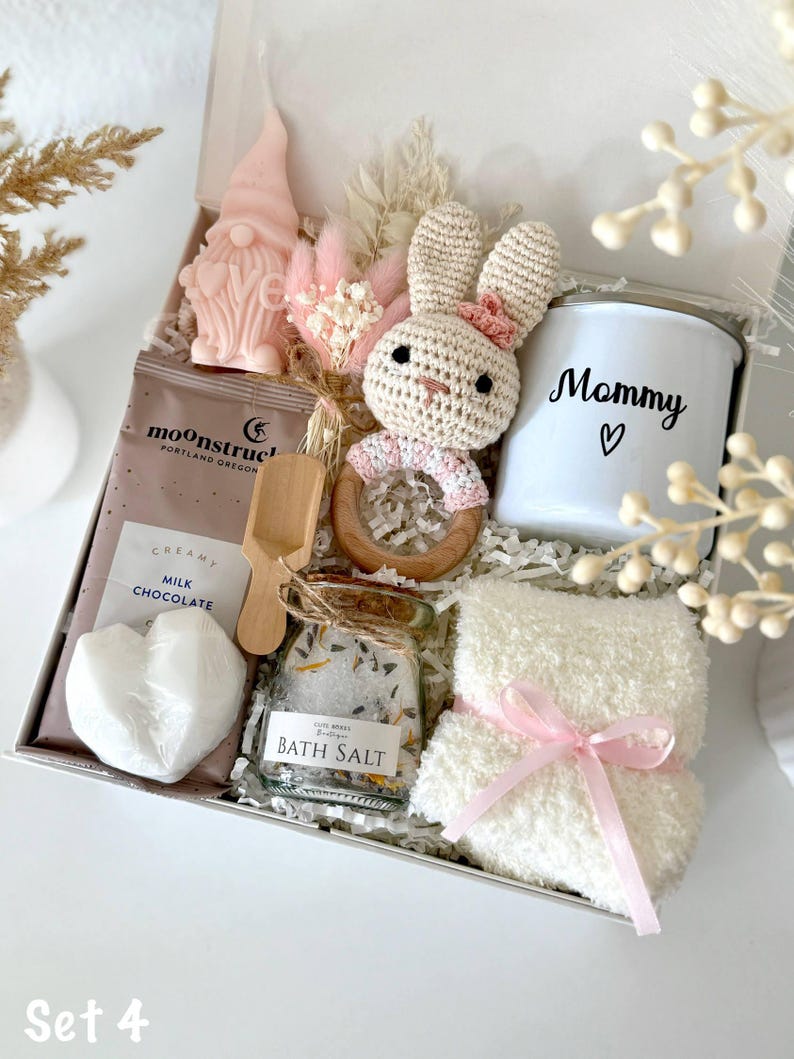 K&ouml;nnte beinhalten: Geschenkbox mit einer rosa Zwergkerze, einem geh&auml;kelten Hasen, einer wei&szlig;en Tasse mit "Mommy" und einem Herz, Badesalz, einer herzf&ouml;rmigen Seife und einem flauschigen wei&szlig;en Waschlappen, der mit einem rosa Band gebunden ist.