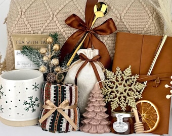 Op de afbeelding: Een cadeaupakket met een beige gebreide deken, een witte mok met groene sneeuwvlokpatronen, een bruin lederen notitieboek met een gouden pen, een boomvormige kaars, een potje honing, een gedroogde sinaasappelschijf en een klein jute zakje.