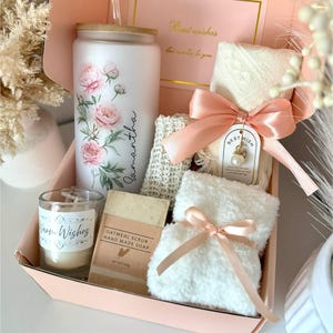 May include: A gift set in a pink box includes a white tumbler with pink floral design and the name "Samantha", a candle, soap, washcloths, and a card that says "Best wishes". The set is tied with a pink ribbon.