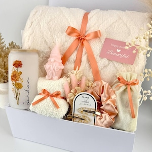 May include: A gift set containing a cream-colored textured blanket tied with a peach ribbon, a frosted glass tumbler with floral art and the name Jordan, a pink gnome candle, a small white towel, a peach scrunchie, hair clips, and a gift bag.