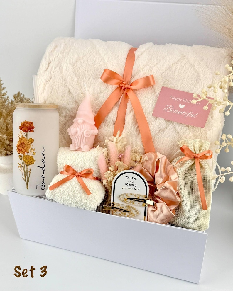 May include: A gift set containing a cream-colored textured blanket tied with a peach ribbon, a frosted glass tumbler with floral art and the name Jordan, a pink gnome candle, a small white towel, a peach scrunchie, hair clips, and a gift bag.