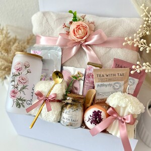 May include: A white gift box filled with self-care items. Includes a rolled white towel with a pink bow and floral accent, a floral-decorated tumbler, bath salts, tea packets, a bath bomb, and a candle. A gold spoon and washcloth are also included.