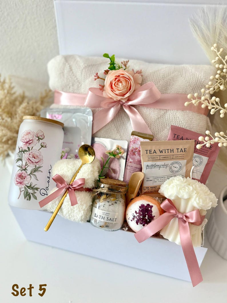 May include: A white gift box filled with self-care items. Includes a rolled white towel with a pink bow and floral accent, a floral-decorated tumbler, bath salts, tea packets, a bath bomb, and a candle. A gold spoon and washcloth are also included.