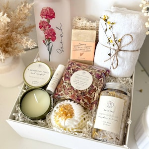 May include: A white gift box containing self-care products. It features a frosted glass with a floral design, a candle, soap, bath salts, a bath bomb, and towels. The glass is inscribed with the name "Sarah".