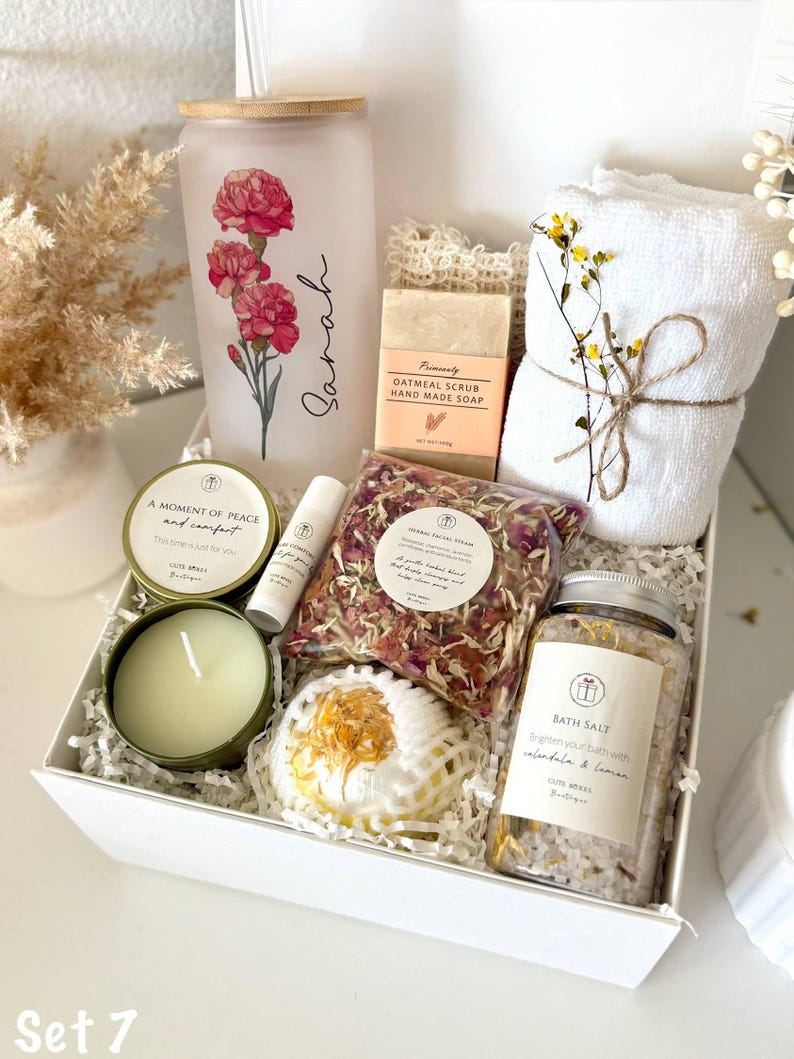 May include: A white gift box containing self-care products. It features a frosted glass with a floral design, a candle, soap, bath salts, a bath bomb, and towels. The glass is inscribed with the name "Sarah".