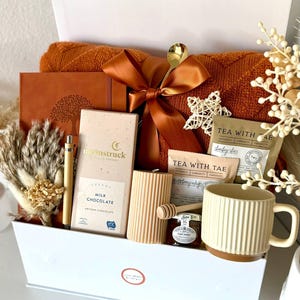 Puede incluir: Una caja de regalo llena de varios art&iacute;culos en tonos c&aacute;lidos. Incluye una manta color &oacute;xido, un diario, chocolate, t&eacute;, una taza, una vela y miel. Una cuchara dorada y un lazo decorativo a&ntilde;aden un toque de elegancia. La caja est&aacute; etiquetada "Set 13".