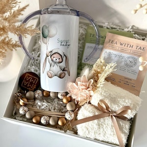 K&ouml;nnte beinhalten: Ein Baby-Geschenkset in einer wei&szlig;en Box. Es enth&auml;lt einen Trinkbecher mit Hasenmotiv und dem Schriftzug "Sweet baby", Teebeutel, einen Schnullerclip und ein Paar wei&szlig;e Socken, die mit einem braunen Band gebunden sind.