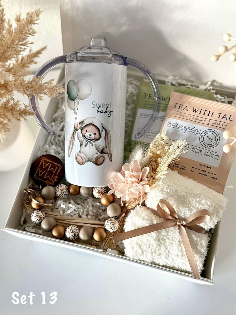 K&ouml;nnte beinhalten: Ein Baby-Geschenkset in einer wei&szlig;en Box. Es enth&auml;lt einen Trinkbecher mit Hasenmotiv und dem Schriftzug "Sweet baby", Teebeutel, einen Schnullerclip und ein Paar wei&szlig;e Socken, die mit einem braunen Band gebunden sind.