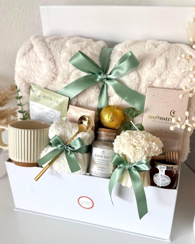 Puede incluir: Una caja de regalo blanca llena de art&iacute;culos de cuidado personal. Incluye una manta esponjosa, taza, t&eacute;, sales de ba&ntilde;o, vela, miel y una cuchara dorada. Los art&iacute;culos est&aacute;n atados con cintas verde salvia, creando un tema de spa.