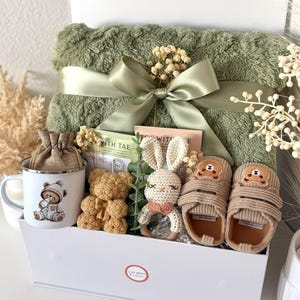 K&ouml;nnte beinhalten: Eine wei&szlig;e Geschenkbox gef&uuml;llt mit Babyartikeln. Enth&auml;lt eine gr&uuml;ne Decke mit Satinband, eine Tasse mit B&auml;renillustration, zwei Teddyb&auml;ren, ein geh&auml;keltes H&auml;schen und Babyschuhe. Die Box ist mit "Set 16" beschriftet.