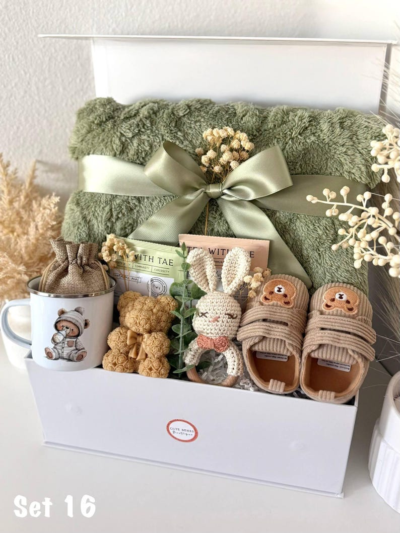 K&ouml;nnte beinhalten: Eine wei&szlig;e Geschenkbox gef&uuml;llt mit Babyartikeln. Enth&auml;lt eine gr&uuml;ne Decke mit Satinband, eine Tasse mit B&auml;renillustration, zwei Teddyb&auml;ren, ein geh&auml;keltes H&auml;schen und Babyschuhe. Die Box ist mit "Set 16" beschriftet.
