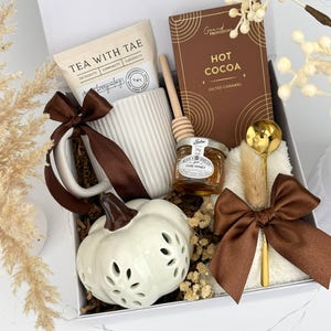 May include: A gift box containing a white ribbed mug with a brown ribbon, a packet of Tea With Tae, a Grand Provisions salted caramel hot cocoa box, a wooden honey dipper, a jar of Wilkin & Sons pure honey, a gold spoon, and a white ceramic pumpkin.