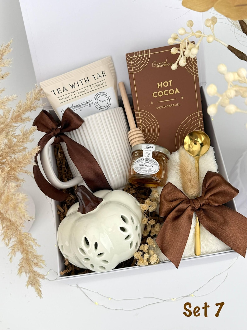May include: A gift box containing a white ribbed mug with a brown ribbon, a packet of Tea With Tae, a Grand Provisions salted caramel hot cocoa box, a wooden honey dipper, a jar of Wilkin & Sons pure honey, a gold spoon, and a white ceramic pumpkin.