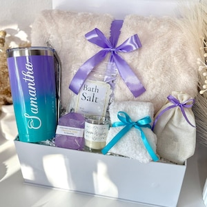 May include: A white gift box filled with self-care items. Includes a plush blanket tied with a purple ribbon, a personalized tumbler, bath salts, a candle, soap, a small towel with a blue ribbon, and a drawstring bag. The tumbler is teal and purple with the name "Samantha".