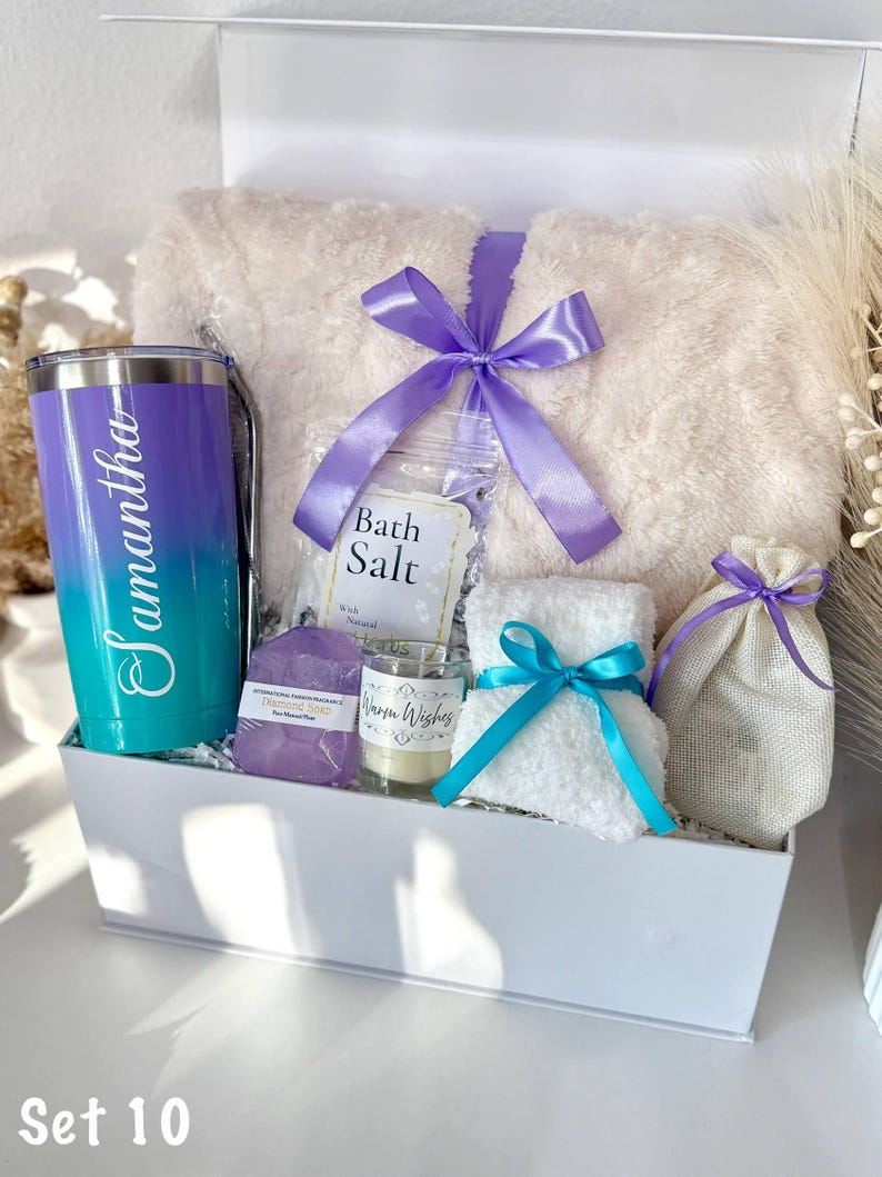 May include: A white gift box filled with self-care items. Includes a plush blanket tied with a purple ribbon, a personalized tumbler, bath salts, a candle, soap, a small towel with a blue ribbon, and a drawstring bag. The tumbler is teal and purple with the name "Samantha".