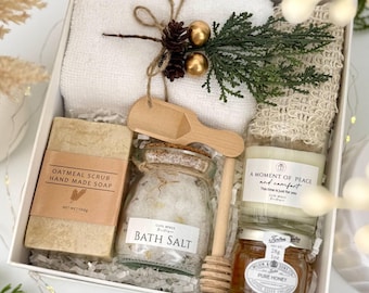 May include: A white gift box containing a white towel, a beige loofah, a bar of oatmeal scrub soap, a glass jar of bath salt, a small wooden scoop, a candle labeled A Moment of Peace and Comfort, and a 1 oz jar of pure honey.