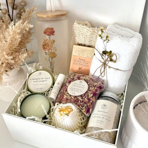 May include: A white gift box filled with self-care items. Includes a frosted glass tumbler with floral design, soap, bath salts, a candle, lip balm, a bath bomb, and a small towel. The text "Set 5" is visible.