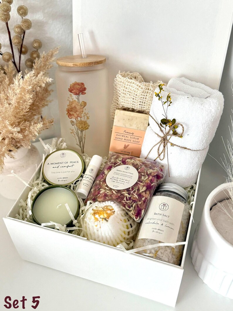 May include: A white gift box filled with self-care items. Includes a frosted glass tumbler with floral design, soap, bath salts, a candle, lip balm, a bath bomb, and a small towel. The text "Set 5" is visible.