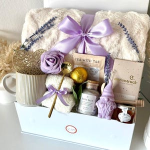 Puede incluir: Una cesta de regalo blanca llena de art&iacute;culos de cuidado personal. Incluye una manta cremosa y esponjosa atada con un lazo lavanda, una taza, t&eacute;, sales de ba&ntilde;o, un jab&oacute;n con forma de gnomo y miel. Ramas de lavanda y una cuchara dorada decoran el conjunto.