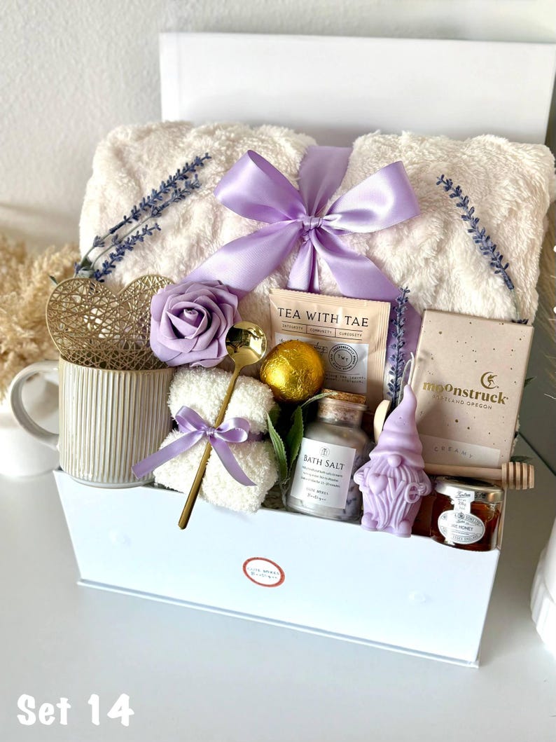Puede incluir: Una cesta de regalo blanca llena de art&iacute;culos de cuidado personal. Incluye una manta cremosa y esponjosa atada con un lazo lavanda, una taza, t&eacute;, sales de ba&ntilde;o, un jab&oacute;n con forma de gnomo y miel. Ramas de lavanda y una cuchara dorada decoran el conjunto.