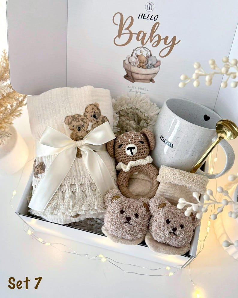 May include: A gift box labeled Hello Baby containing a cream-colored muslin blanket with bear embroidery and fringe, a crocheted bear rattle, a speckled ceramic mug with the word mom, a gold-toned spoon, and a pair of fuzzy bear-faced baby booties.