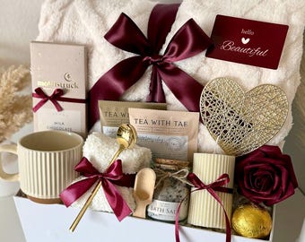 May include: A white gift box filled with self-care items. Includes a fluffy white blanket, a mug, chocolate, tea, bath salts, a candle, a gold heart, a rose, and a card that says "hello Beautiful". All items are tied with burgundy ribbons.