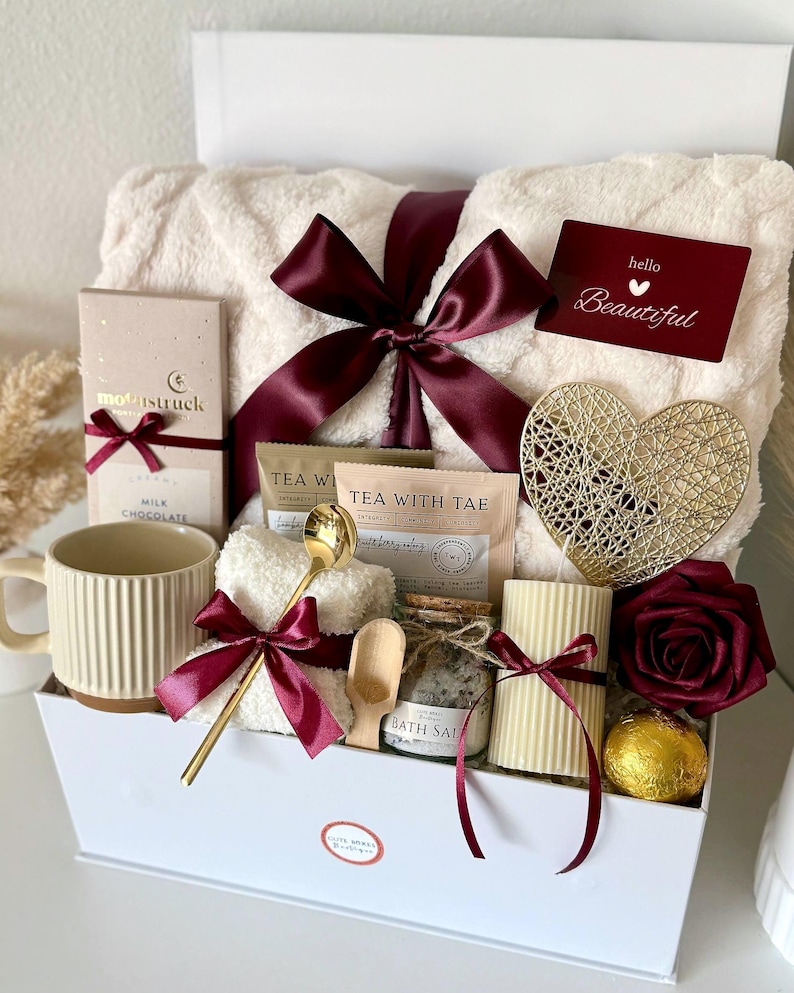 K&ouml;nnte beinhalten: Eine wei&szlig;e Geschenkbox mit Pflegeprodukten. Enth&auml;lt eine flauschige wei&szlig;e Decke, eine Tasse, Schokolade, Tee, Badesalz, eine Kerze, ein goldenes Herz, eine Rose und eine Karte mit "hello Beautiful". Burgunderfarbene B&auml;nder.