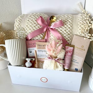 Puede incluir: Una caja de regalo blanca llena de una manta de punto color crema, cinta rosa, cuchara dorada, taza, t&eacute;, miel, chocolate y crema de manos. La caja est&aacute; etiquetada como "Set 5".