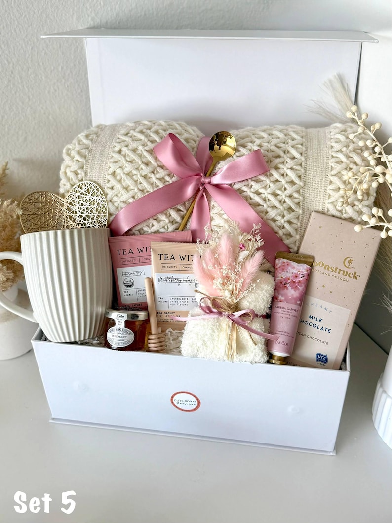 Puede incluir: Una caja de regalo blanca llena de una manta de punto color crema, cinta rosa, cuchara dorada, taza, t&eacute;, miel, chocolate y crema de manos. La caja est&aacute; etiquetada como "Set 5".