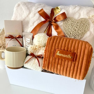 May include: A gift set in a white box containing a cream-colored textured blanket with a brown ribbon, a gold heart ornament, a ribbed mug, a chocolate bar, a small white flower candle, and a rust-colored corduroy pouch with the name Samantha.