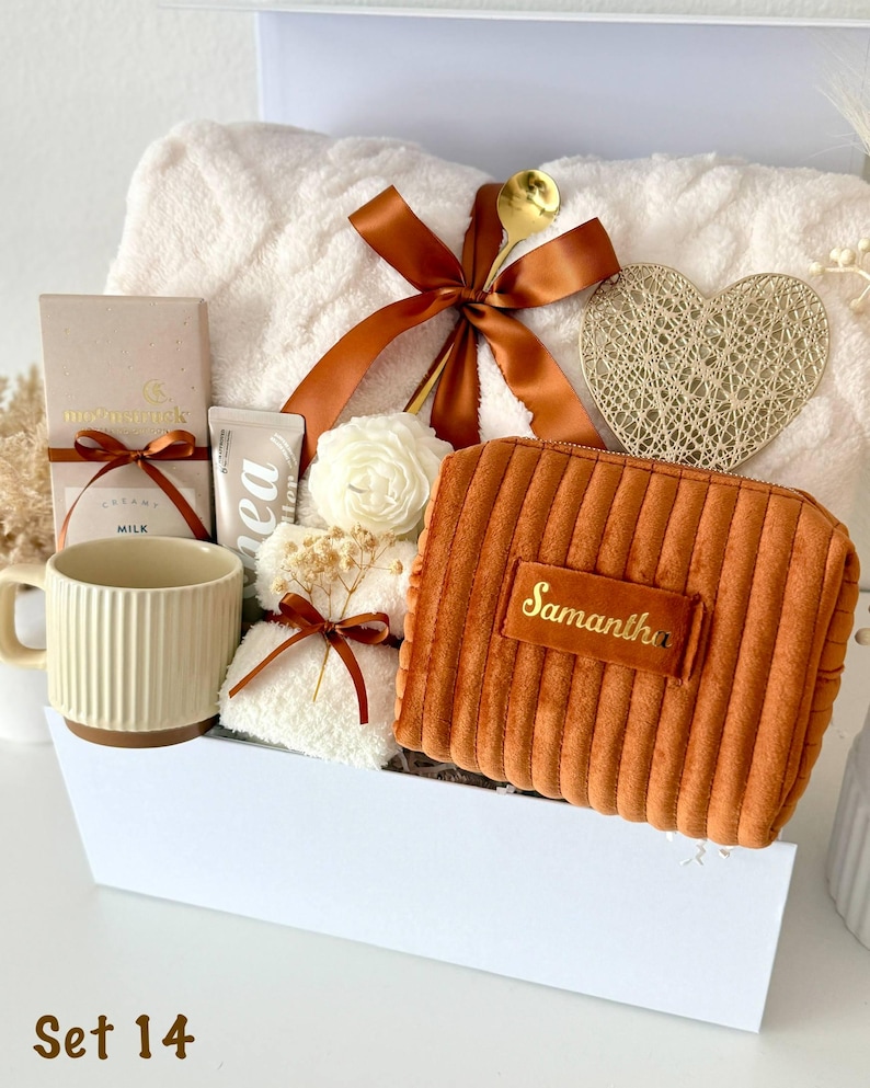 May include: A gift set in a white box containing a cream-colored textured blanket with a brown ribbon, a gold heart ornament, a ribbed mug, a chocolate bar, a small white flower candle, and a rust-colored corduroy pouch with the name Samantha.